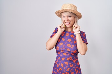 Young caucasian woman wearing flowers dress and summer hat smiling pulling ears with fingers, funny gesture. audition problem