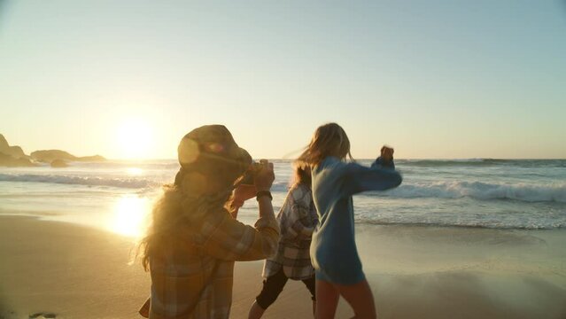Three young women hang out at beach at sunset. Happy smiling friends run around barefoot in cinematic location near ocean. Photographer make photos of friends