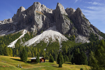 Panorama of Dolomites in Italy with yellow-green fir-trees around them. Sunny day. The brown hut is...