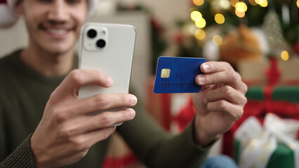 Young hispanic man using smartphone and credit card sitting on floor by christmas tree at home