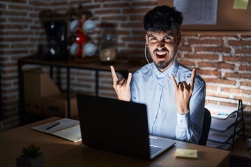 Young hispanic man with beard working at the office at night shouting with crazy expression doing rock symbol with hands up. music star. heavy concept.