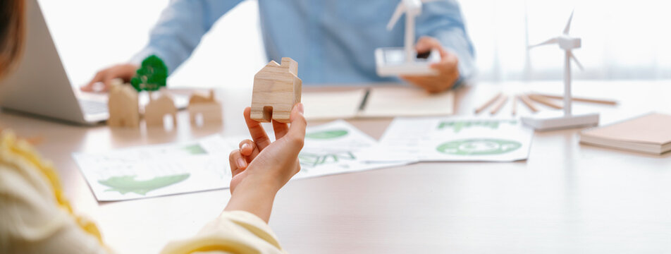 Cropped image of businesswoman presents eco-friendly house by using green design to manager while holding the wooden house block at table with windmill model placed with document. Delineation.