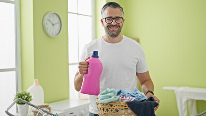 Grey-haired man smiling confident holding detergent bottle at laundry room