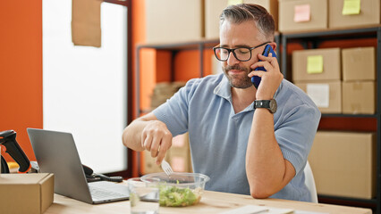 Grey-haired man ecommerce business talking on smartphone eating salad at office