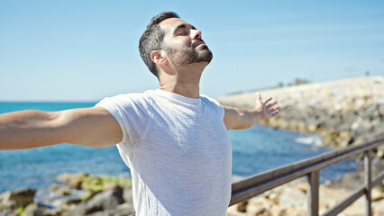 Young hispanic man breathing with closed eyes and arms open at seaside - obrazy, fototapety, plakaty