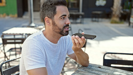 Young hispanic man sending voice message by smartphone sitting on table at coffee shop terrace