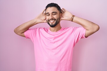 Hispanic young man standing over pink background smiling cheerful playing peek a boo with hands...