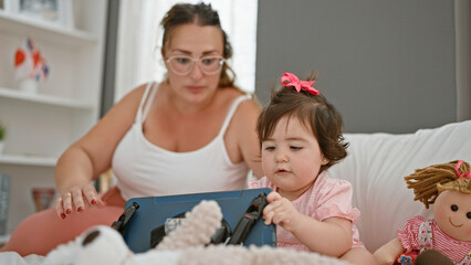 Cosy morning time, mother and daughter, comfortable in pyjamas, sitting together on bed, absorbed in touchpad technology, their beloved gadget, in a homely bedroom room