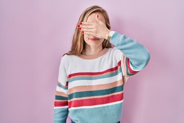 Young blonde woman standing over pink background covering eyes with hand, looking serious and sad....
