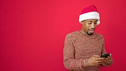 African american man wearing christmas hat using smartphone smiling over isolated red background