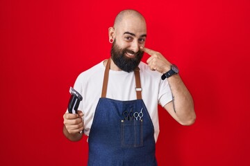 Naklejka premium Young hispanic man with beard and tattoos wearing barber apron holding razor pointing with hand finger to face and nose, smiling cheerful. beauty concept
