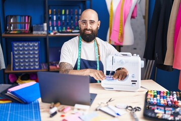 Young bald man tailor using sewing machine and laptop at clothing factory