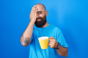 Young hispanic man with beard and tattoos drinking a cup of coffee covering one eye with hand,...