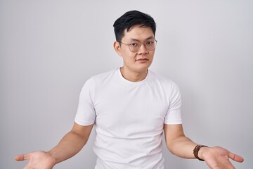 Young asian man standing over white background clueless and confused with open arms, no idea...