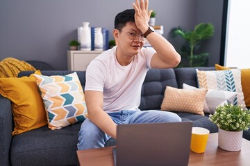 Young asian man using laptop at home sitting on the sofa surprised with hand on head for mistake,...
