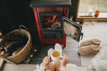 Young woman sitting by the fireplace in white sweater, drinking tea in cozy log cabin.
