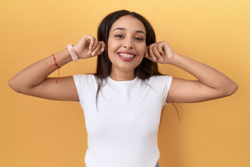 Young arab woman wearing casual white t shirt over yellow background smiling pulling ears with...