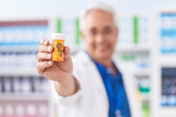 Middle age grey-haired man pharmacist smiling confident holding pills bottle at pharmacy