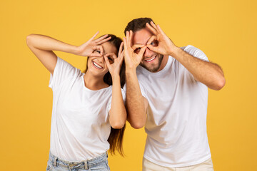 Cheerful young european man and woman in white t-shirts making fun glasses gesture with hands