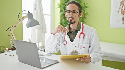 Obraz premium Young hispanic man doctor using laptop writing on clipboard at clinic