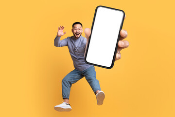 Asian man holding large cellphone blank screen leaping in studio