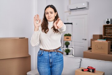 Young hispanic woman holding keys of new home pointing with finger to the camera and to you, confident gesture looking serious