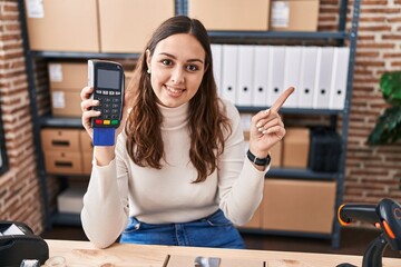 Fototapeta na wymiar Young hispanic woman working at small business ecommerce holding dataphone smiling happy pointing with hand and finger to the side
