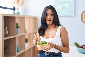 Brunette woman eating a salad at home in shock face, looking skeptical and sarcastic, surprised with open mouth