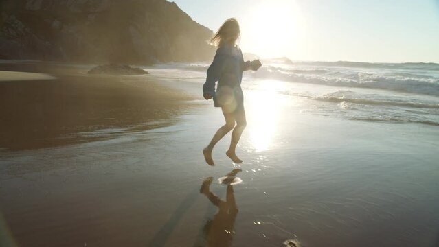 Happy young woman in oversized sweater run barefoot on beach in ocean surf waves. Cinematic moment of happiness and joy on warm summer evening