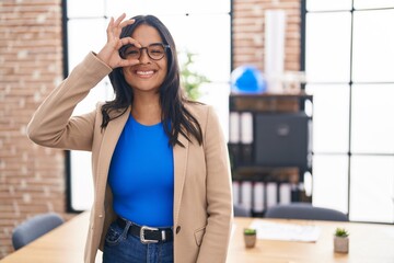 Brunette woman working at the office wearing glasses smiling happy doing ok sign with hand on eye looking through fingers