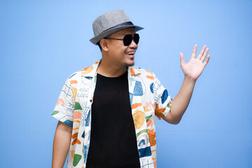 asian man wearing beach shirt pointing a finger to vacation discount offer. Isolated on blue background. Vacation concept