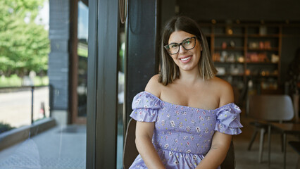 Joyful brunette hispanic woman sitting at cafe table, enjoying an indoors coffee. beautifully casual, she's confidently smiling, positively radiating happiness through her glasses.