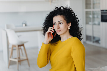 Curly Spanish young woman in yellow sweater talking by phone remote working at home on cozy sofa....