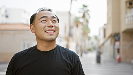 Joyful young chinese man, confidently smiling, looking to the sky while standing in the urban street