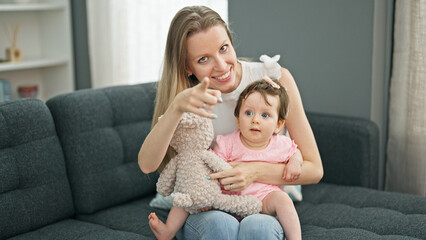 Mother and daughter sitting on sofa playing with teddy bear looking to the camera at home