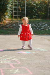 A cute little girl toddler in a red dress walks on the asphalt with chalk drawings