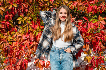 Portrait of a beautiful girl in a shirt walking in the park against the background of a bush with red autumn leaves, a cozy autumn atmosphere