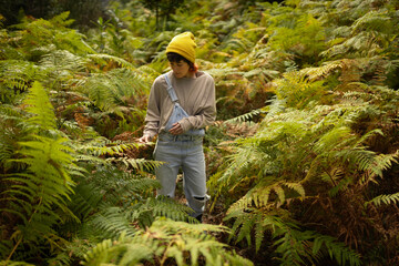 Woman with mobile phone and headphones in fern forest wearing yellow hat, listening to music,...