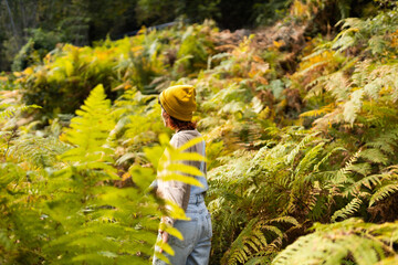 Woman with mobile phone and headphones in fern forest wearing yellow hat, listening to music,...