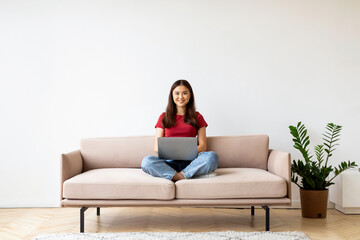 Happy young asian woman with laptop sitting cross-legged on sofa at home