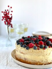 Homemade vanilla cake with buttercream frosting and fresh berries on a white surface with glasses of white wine in the background. Christmas cake. Copy space