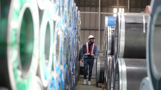 Warehouse Inspection: A meticulous warehouse worker, clad in safety gear, conducts a thorough inspection of stacked metal coils, ensuring operational efficiency and quality control.
