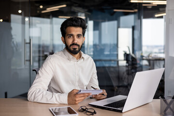 Fototapeta premium Portrait of an Indian young businessman working in the office at a table with a laptop, holding a notebook, looking seriously at the camera