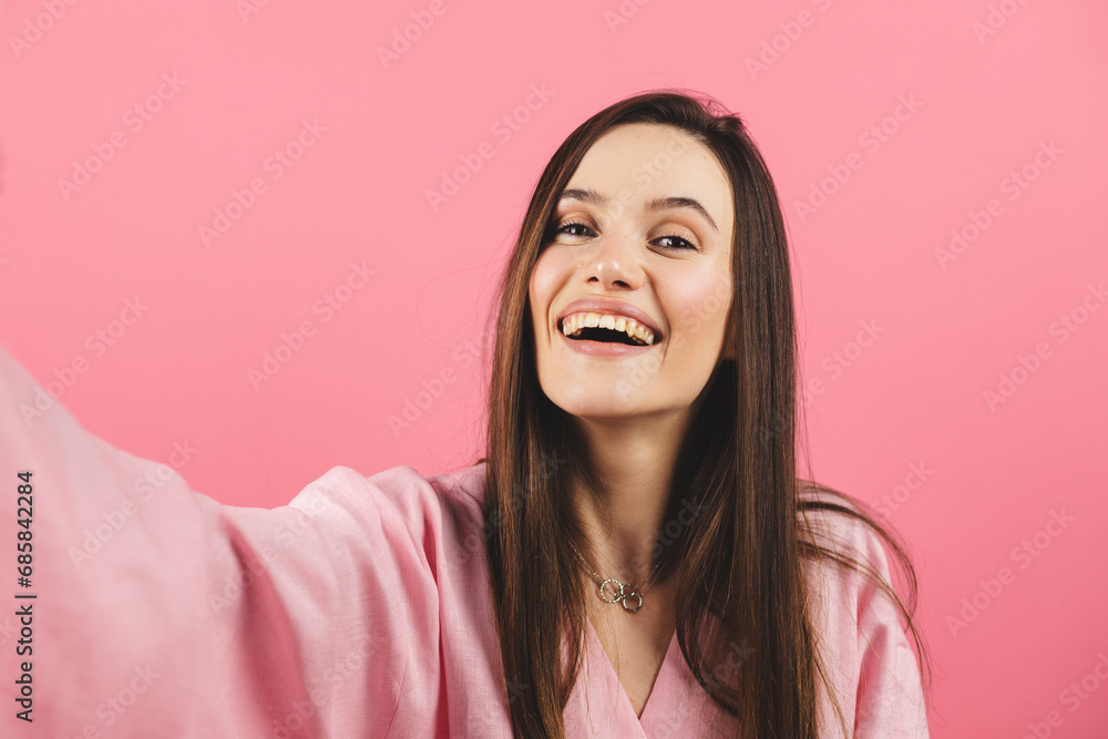 Wall mural Young smiling brunette woman wear pink dress doing selfie shot on mobile phone post photo on social network isolated on pink background. Woman make video call, selfie on her mobile phone.