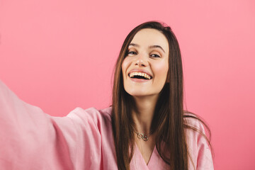 Young smiling brunette woman wear pink dress doing selfie shot on mobile phone post photo on social network isolated on pink background. Woman make video call, selfie on her mobile phone.