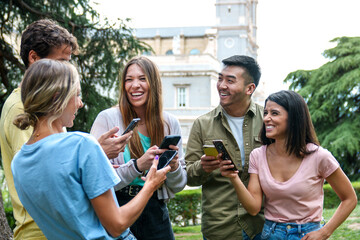 Five joyful friends sharing a laugh outside while checking their phones, with trees and architecture in the background.