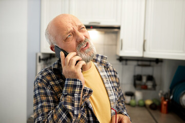 Senior man talking on phone standing in kitchen in early morning, ordering goods or home repair services, looking up with puzzled facial expression, giving answer to some questions of his interlocutor