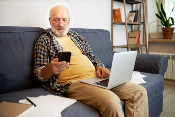 Fototapeta premium Aged man in glasses with big belly sitting on sofa full of paper documents with laptop on knees, holding smartphone, working or paying taxes and utility bills using mobile application