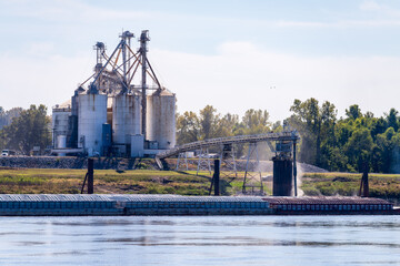 Tugboat pushing freight barges past grain loading dock in farming country in Kentucky