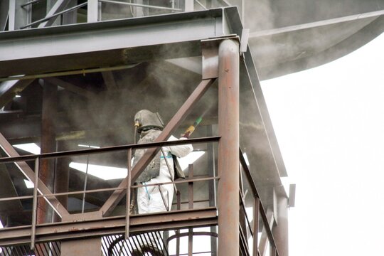 Sand blasting process. Industrial worker in protective uniform cleaning surface of big steel construction before painting on high altitude.